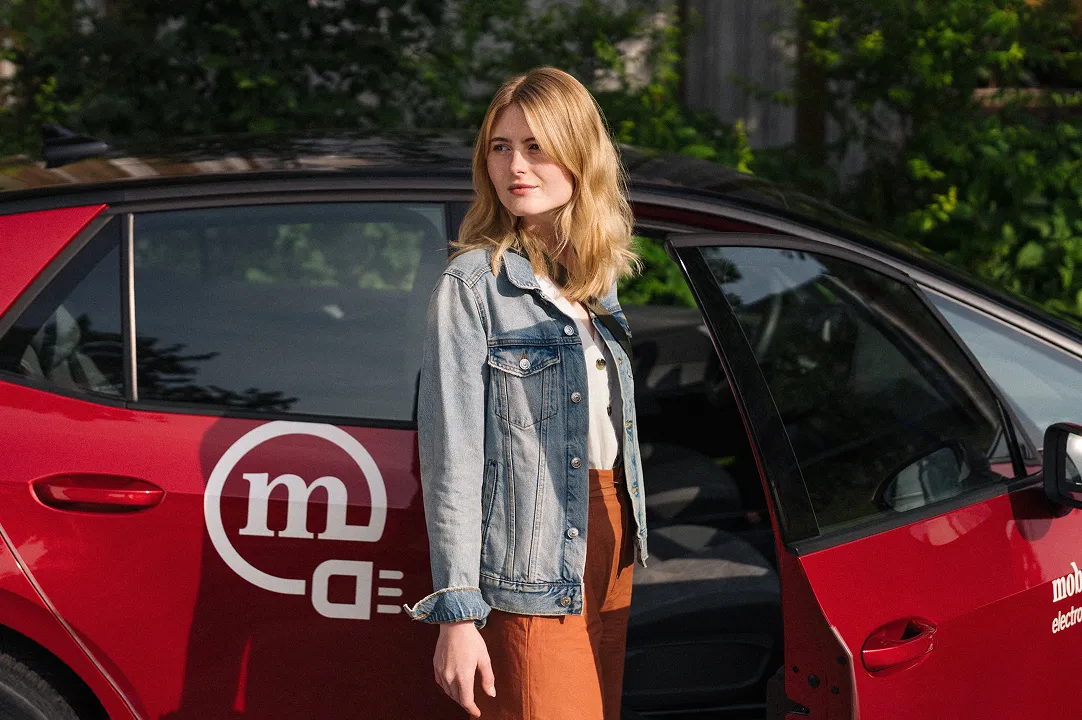 Une femme se tient à côté d’une voiture rouge de car sharing Mobility, avec la porte ouverte. Elle utilise la mobilité partagée au quotidien.