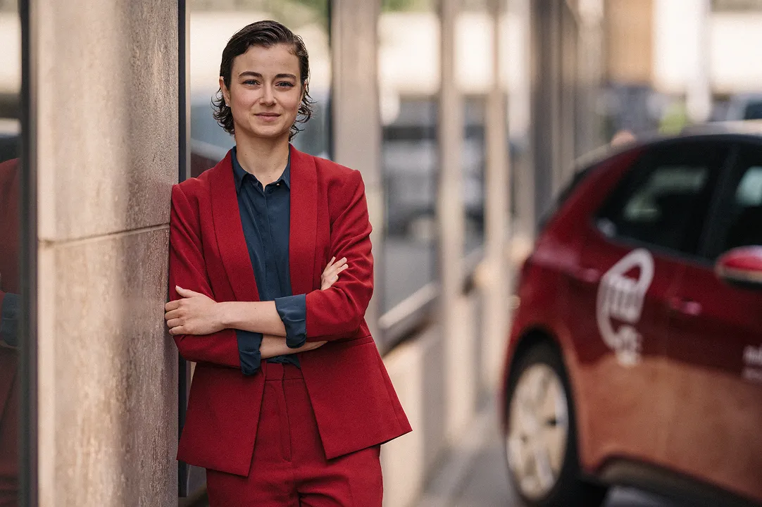 Une femme en tailleur rouge appuyée contre le mur d’un bâtiment en ville, à côté d’un véhicule de car sharing Mobility.