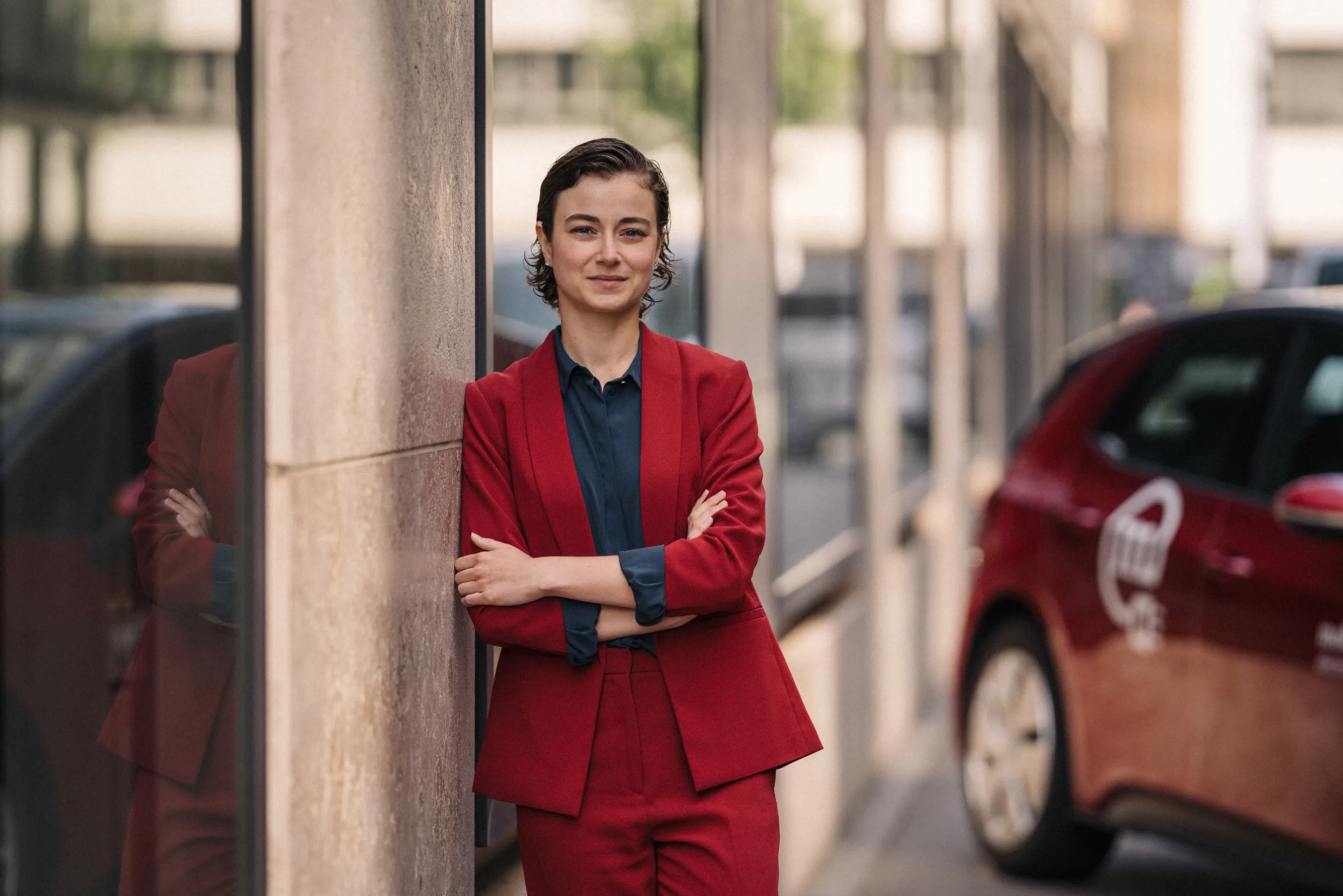 Une femme en tailleur rouge appuyée contre le mur d’un bâtiment en ville, à côté d’un véhicule de car sharing Mobility.