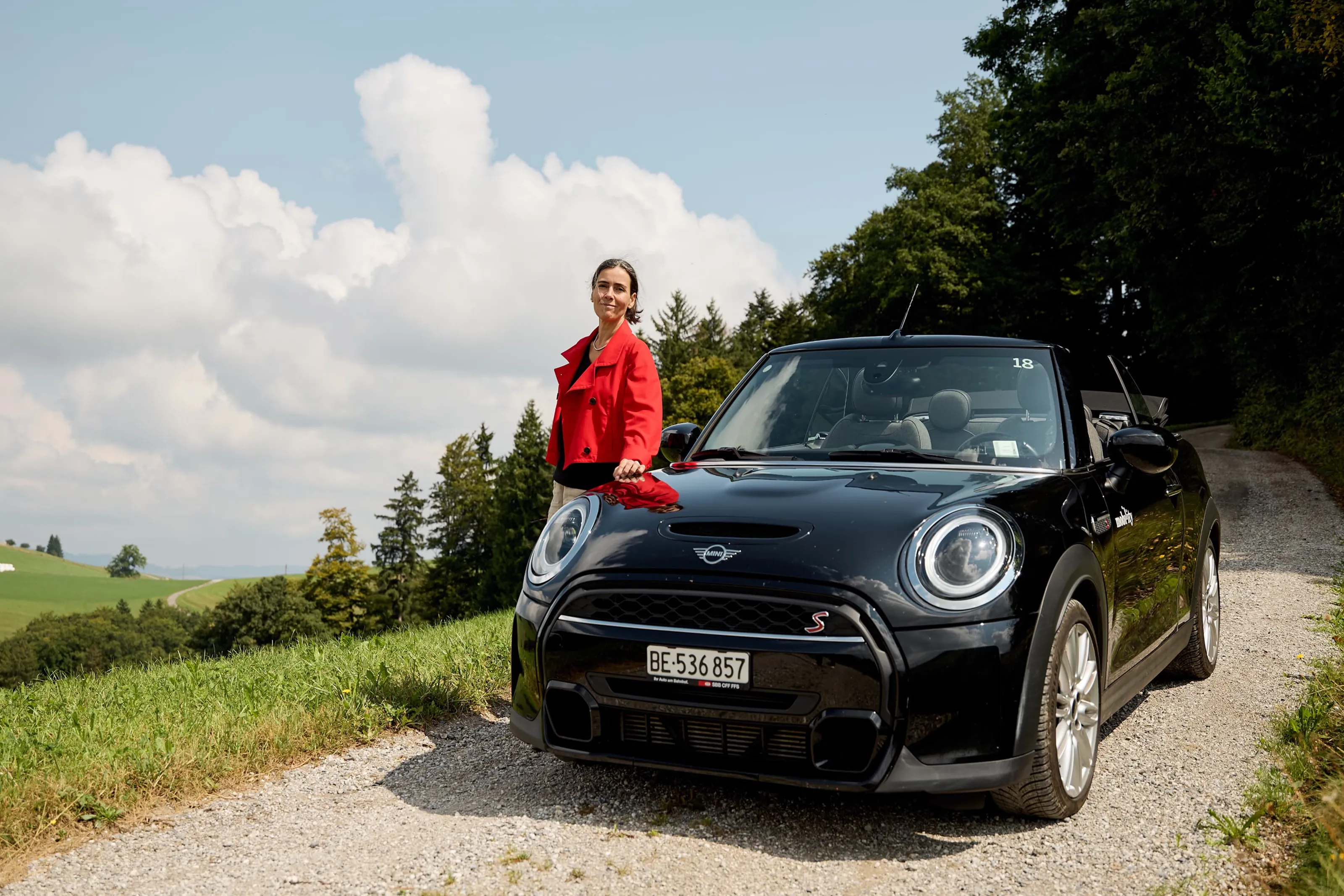 Une femme près d’un cabriolet noir, sur une route de campagne avec une vue sur des prés et des arbres.