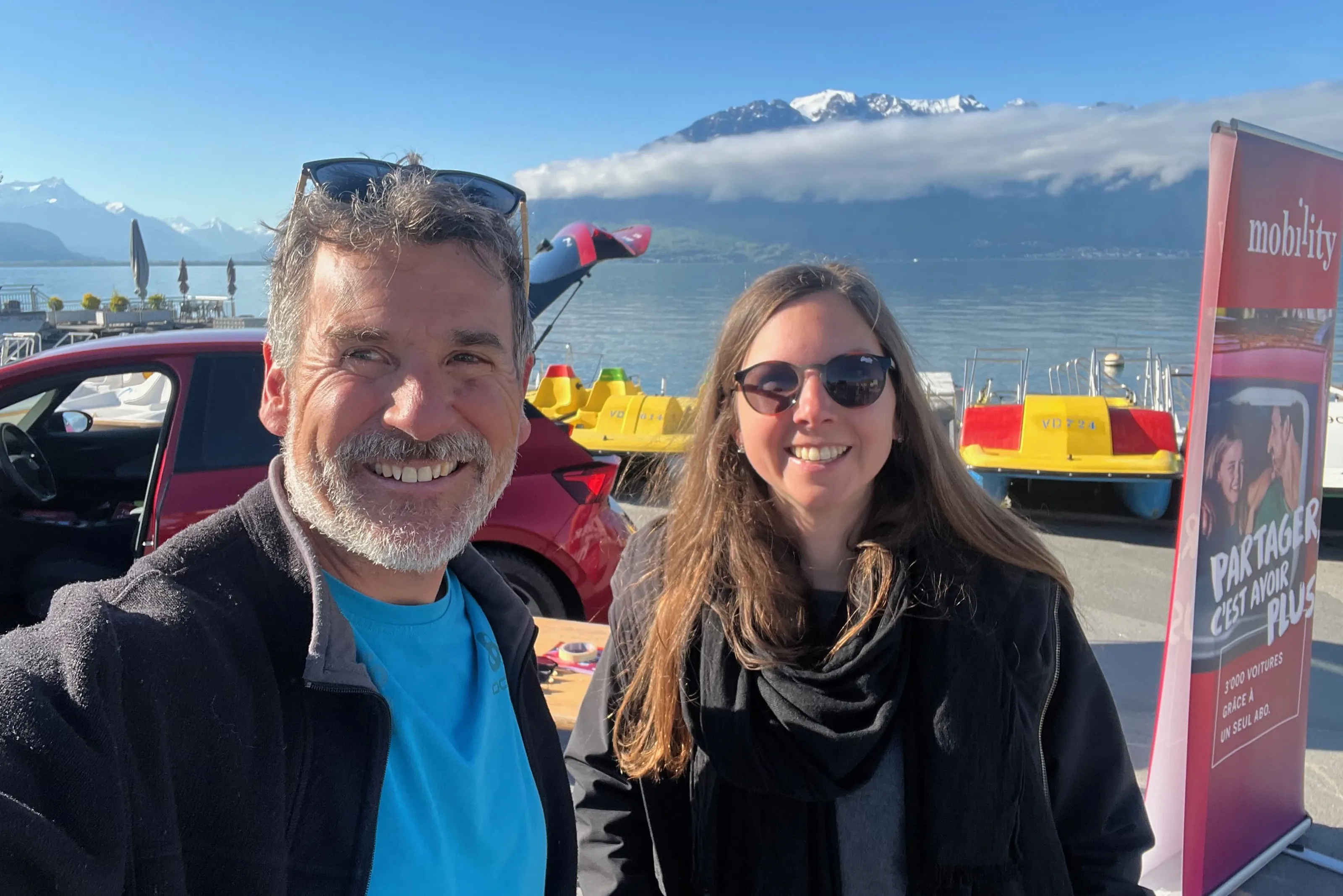 Deux personnes font un selfie au bord d’un lac, devant des montages, près d’une voiture de car sharing rouge et de pédalos.