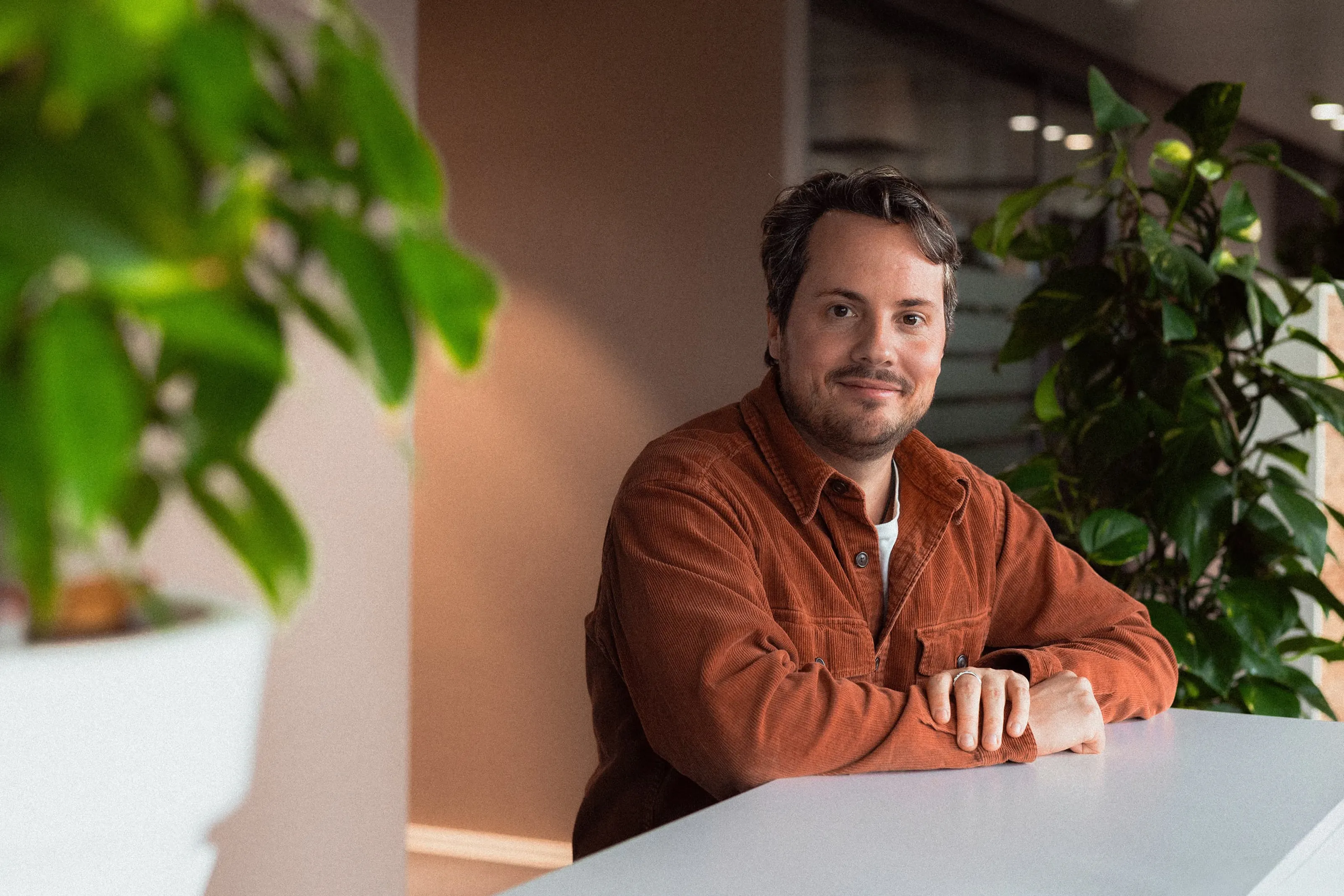 Un homme souriant en chemise marron, assis dans un bureau moderne avec des plantes.