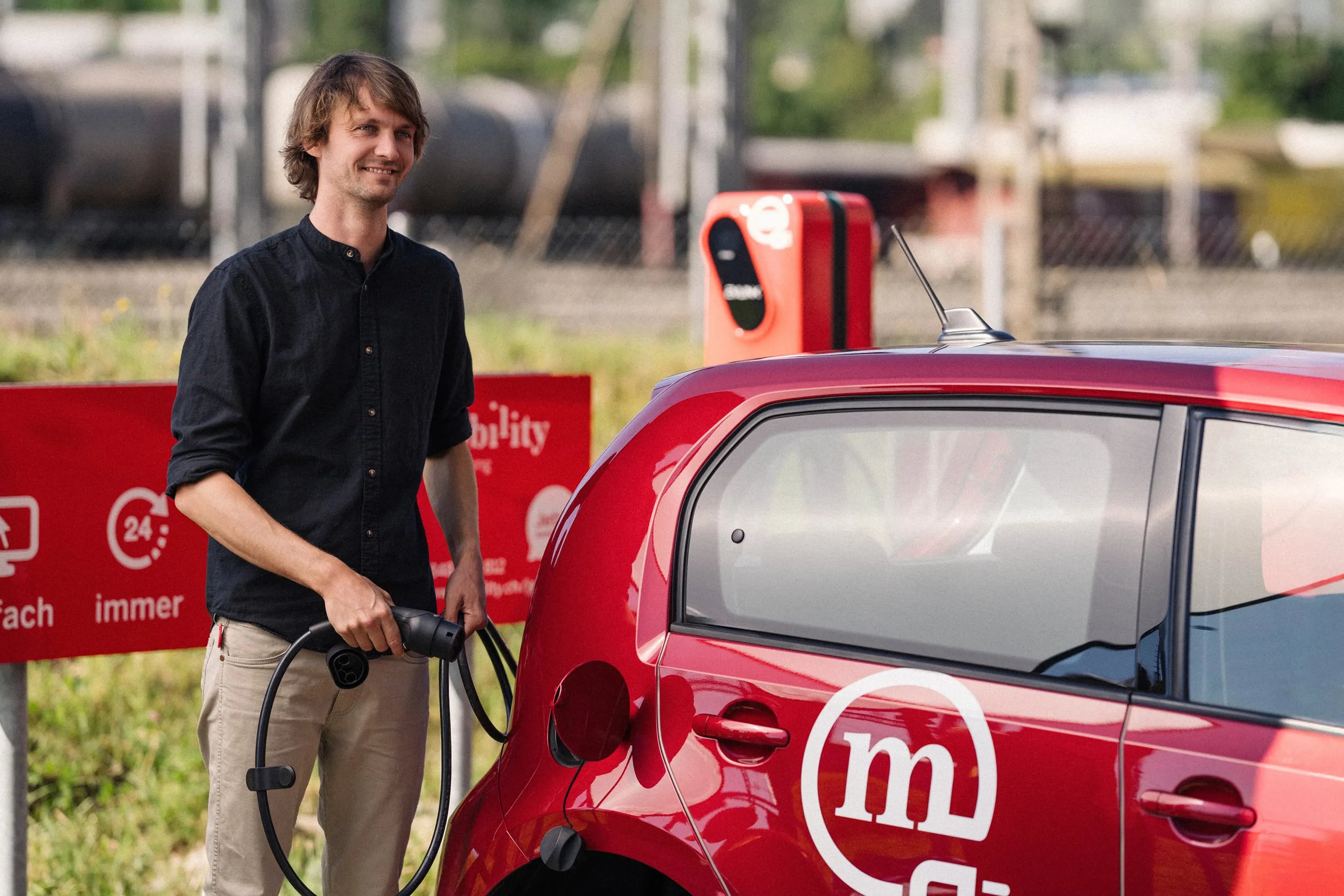 Un homme recharge une voiture électrique rouge de car sharing Mobility à une borne de recharge publique et sourit.